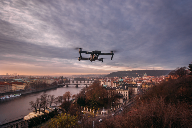 Ein Drohne fliegt über eine Stadtlandschaft mit Gebäuden, Bäumen und Fahrzeugen, einem Fluss und einer Brücke im Hintergrund, unter einem klaren Himmel.