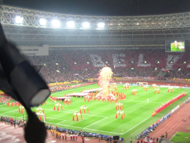 Ein Stadion mit Flutlicht, Eisen Gittern, Zuschauern, stehenden Personen, Werbetafeln, Bildschirmen, einer Überwachungskamera, Fahnen und Dekorationselementen.