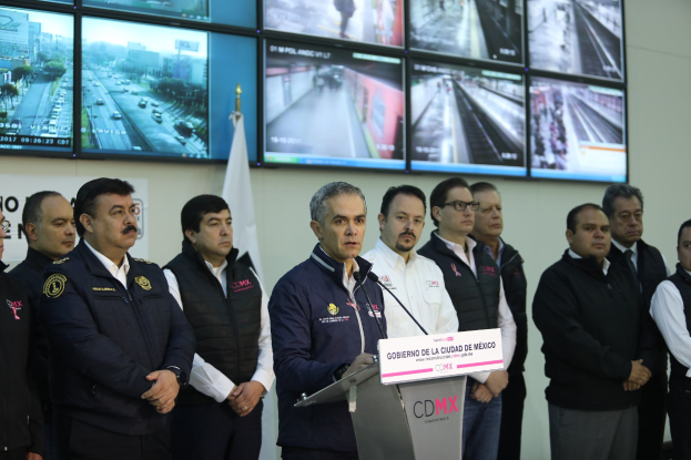 Eine Gruppe von Männern steht vor einem Rednerpult mit einem Mikrofon und einer Tafel mit der Aufschrift "Mexikanische Zoll- und Grenzschutzbehörde", mit Fernsehgeräten und einer Flagge an der Wand dahinter.