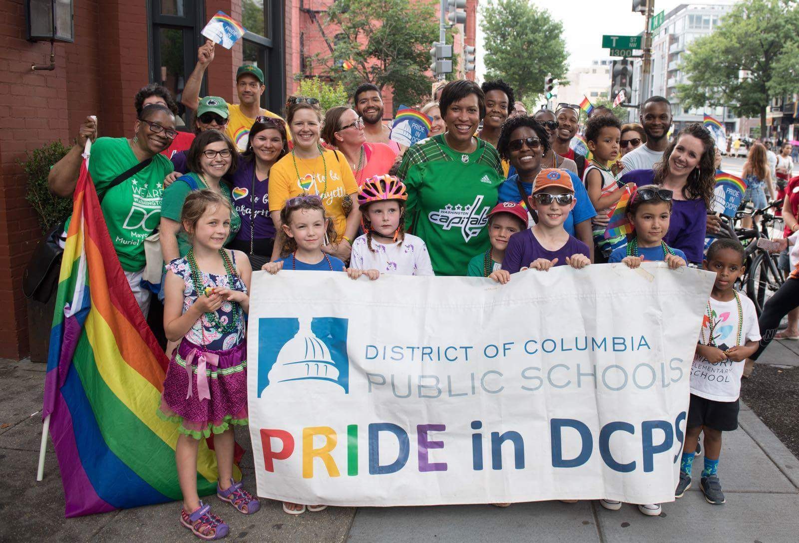 Gruppe von Menschen, die draußen mit einem Banner stehen, auf dem "Stadtteil Washington D.C. Public Schools Pride in DCPS" steht, Kinder mit einer Fahne, ein Radfahrer, Bäume, Gebäude, Straßenschilder und eine bewölkte Himmel.