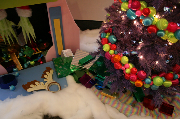Eine Indoor-Szene mit einem geschmückten Weihnachtsbaum auf der rechten Seite, Geschenkboxen und Dekorationen in der Mitte und eine kleine Hütte mit Dekorationen oben links in der Ecke, mit Watte und Wolle am Boden.