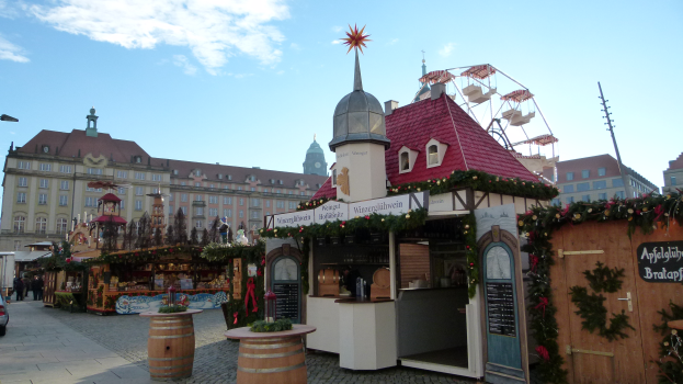 Ein lebendiger Weihnachtsmarkt auf einem Stadtplatz, umgeben von Gebäuden, Straßenlaternen und geschmückten Bäumen, mit Menschen und Fahrzeugen auf der Straße unter einem bewölkten Himmel.