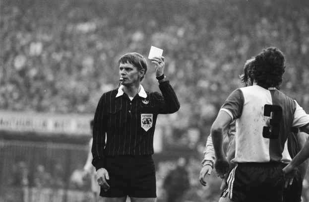 Schwarzes und weißes Bild eines Schiedsrichters mit roter Karte, mit einer Menge von Menschen in Fußballuniformen im Hintergrund.