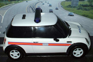 Weißer Polizeiwagen mit "Polizei" Aufschrift auf einer Straße außerhalb der Stadt, umgeben von Verkehr, Gras und Bäumen.