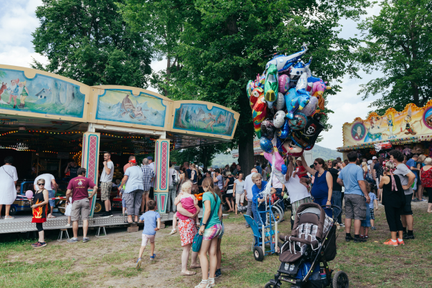 Eine Menschenmenge steht um ein buntes, beleuchtetes Karussell auf einem Volksfest, einige halten Kinderwagen, mit Bäumen und einem bewölktem Himmel im Hintergrund.