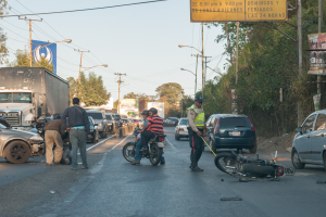 Eine Gruppe von Menschen um ein verunglücktes Motorrad auf der Straße herumstehend mit mehreren Fahrzeugen, einschließlich eines Lastwagens, und einem Hintergrund aus Bäumen, Polen, Lichtern und Brettern unter dem Himmel.