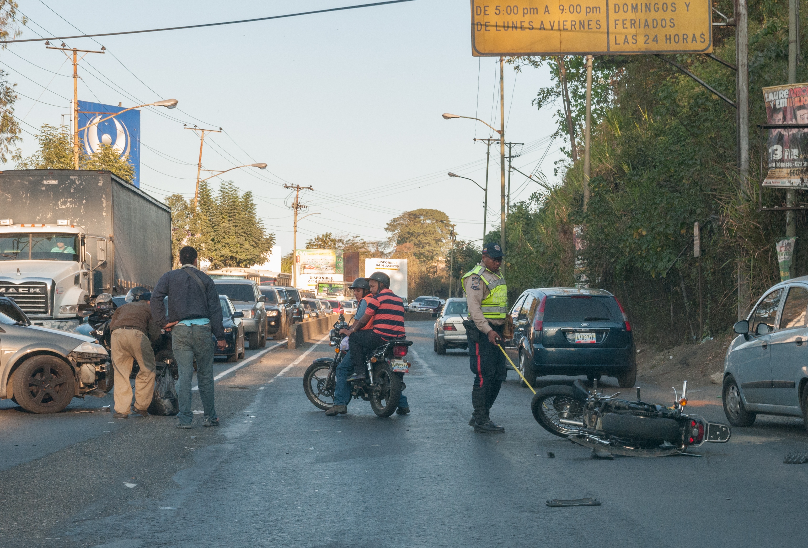 Eine Gruppe von Menschen um ein verunglücktes Motorrad auf der Straße herumstehend mit mehreren Fahrzeugen, einschließlich eines Lastwagens, und einem Hintergrund aus Bäumen, Polen, Lichtern und Brettern unter dem Himmel.