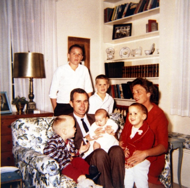 Familie von fünf Personen auf einer Couch im Wohnzimmer sitzend, wobei ein Mann ein Baby hällt, eine Frau mit zwei Kindern und zwei Männer dahinter stehen.