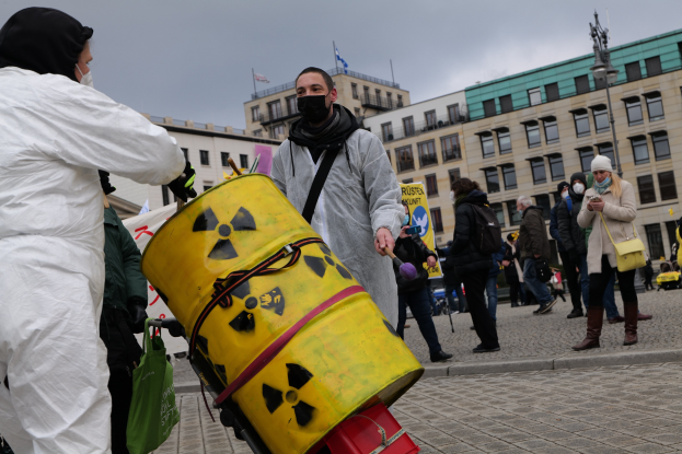Ein Mann in einem weißen Anzug trägt einen gelben Bottich mit einem radioaktiven Symbol, umgeben von maskierten Individuen mit Taschen, mit Gebäuden, Flaggen und einem bewölkten Himmel im Hintergrund.