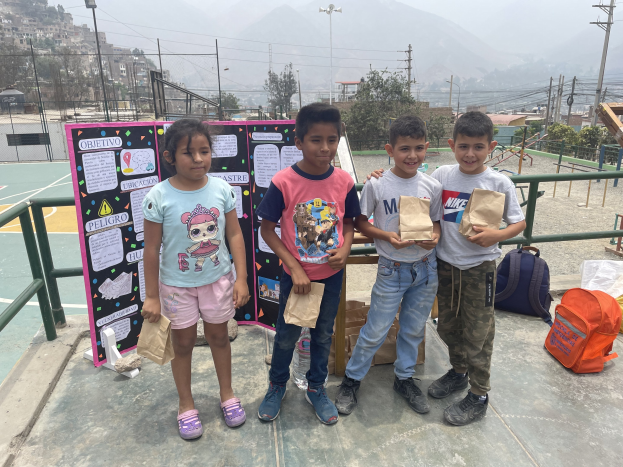 Gruppe von Kindern auf einem Basketballfeld mit Papierbeuteln in den Händen, mit Gegenständen auf dem Boden, einer Tafel mit Text auf der linken Seite und einem Hintergrund aus Zäunen, Pfosten, Straßenlaternen, Bäumen, Gebäuden, Hügeln und Himmel.