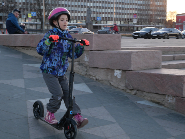 Ein junger Junge in Helm und Handschuhen fährt mit einem Roller eine Treppe hinunter, vorbei an Fahrzeugen, Menschen, Bäumen, Pfosten, Brettern, Gebäuden und einem klaren blauen Himmel.