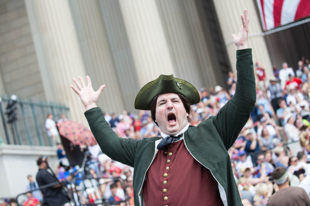 Ein Mann mit Schützendar und grünem Jackett singt vor einer Menge mit Musikinstrumenten, mit einem Geländer im Vordergrund, einem Gebäude im Hintergrund und einer Flagge rechts.