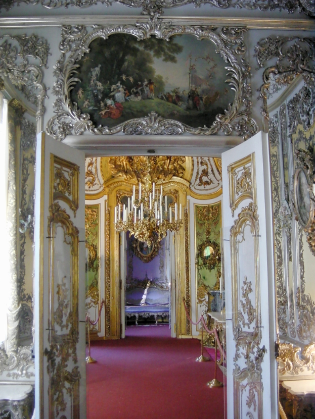 Kerzen auf einem Kronleuchter, Türen mit Gemälden verziert und ein roter Teppich auf dem Boden.