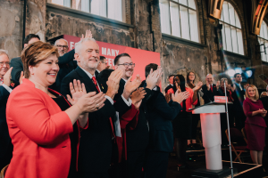 Eine Gruppe von Menschen, die vor einem Publikum applaudieren, mit einem Podium, einem Mikrofon und einer Texttafel rechts, sowie Stühlen, einer Fahne, einer Wand, Fenstern und Lichtern im Hintergrund.