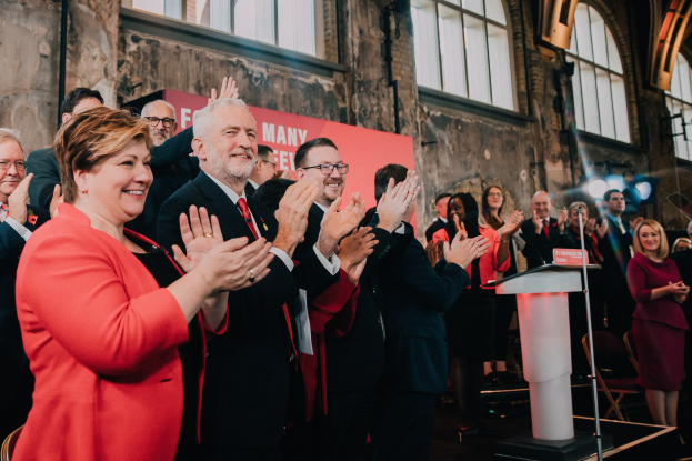 Eine Gruppe von Menschen, die vor einem Publikum applaudieren, mit einem Podium, einem Mikrofon und einer Texttafel rechts, sowie Stühlen, einer Fahne, einer Wand, Fenstern und Lichtern im Hintergrund.