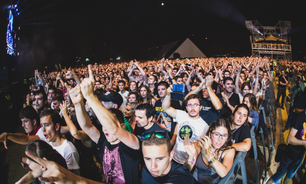 Menschenmenge auf einem Musikfestival mit erhobenen Händen, einige sitzen auf Stühlen und andere stehen, begrenzt von einem Metallzaun auf der rechten Seite, mit Gebäuden und Lichtern im Hintergrund unter einem dunklen Himmel.