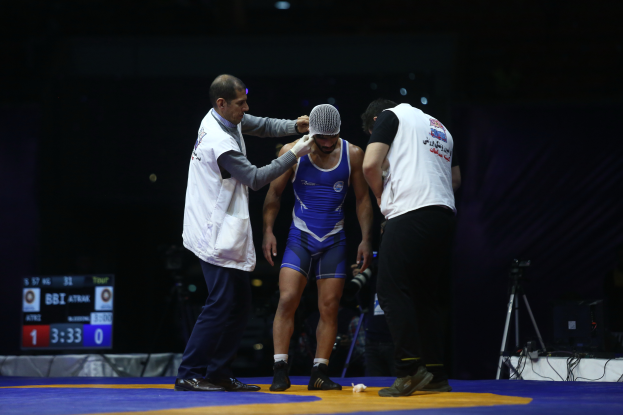 Ein Mann in einem blauen Wrestling-Anzug steht auf einer Wrestling-Matte, umgeben von ein paar Menschen, mit einer Person, die eine Kamera mit Stativ und andere Ausrüstung im Hintergrund hält, und einer digitalen Anzeigetafel mit Zahlen und Text, alles vor einem dunklen Nacht-Hintergrund.