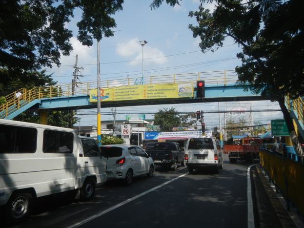 Eine belebte Straße mit Fahrzeugen, eine Brücke mit Geländern und Treppen, Laternen, Verkehrsampeln, Schilder, Bäume, Gebäude und ein bewölktes Himmel.