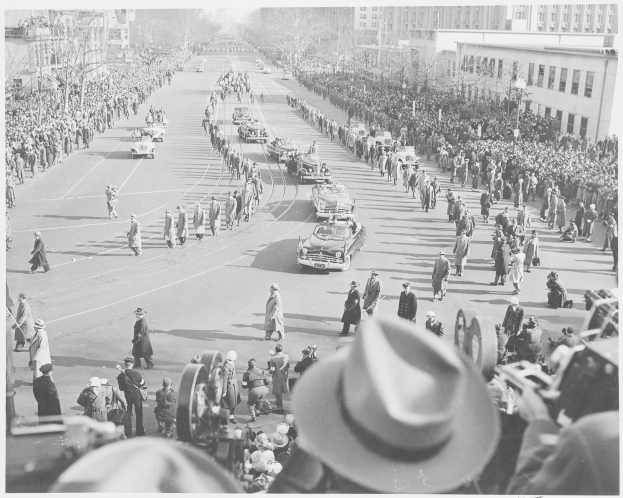 Schwarzes und weißes Foto einer Parade mit Autos und Menschen auf der Straße, einige tragen Hüte, vor einer Kulisse aus Bäumen und Gebäuden.