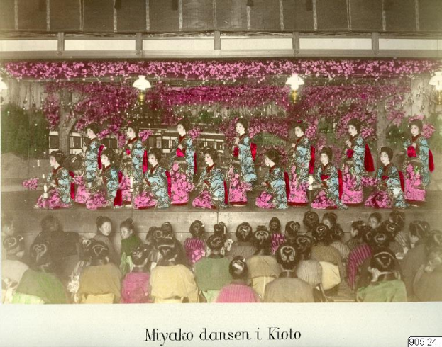 Eine Gruppe von Frauen in bunten Kimonos sitzt auf einer Bühne, die mit Blumen und Lichtern geschmückt ist, vor einem Publikum.