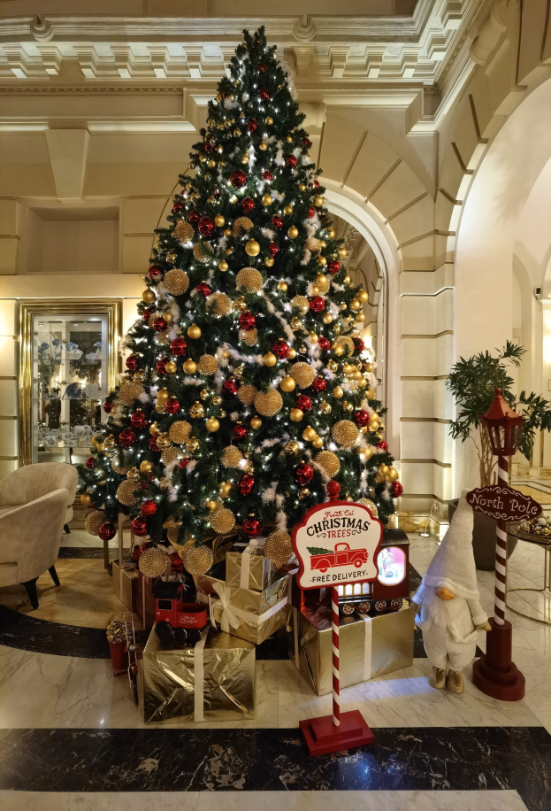 Großer Weihnachtsbaum mit Schmuck und Lichtern im Hotel-Foyer, umgeben von Geschenkboxen, einem Spielzeug, einer Tafel, Mobiliar und einer Pflanze.