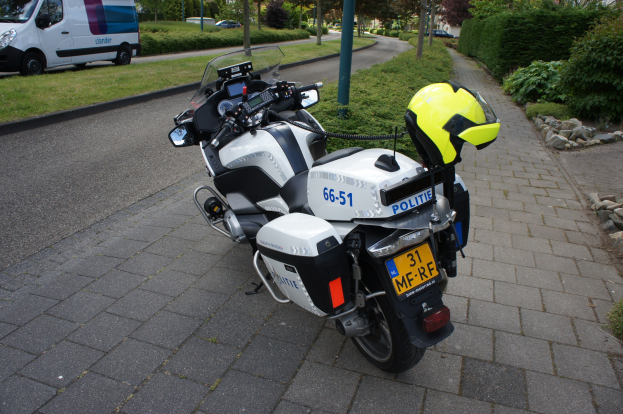 Ein polizeiliches Motorrad am Straßenrand geparkt mit einem Helm darauf, umgeben von Bäumen, Gebäuden und anderen Fahrzeugen.