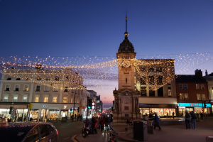Eine belebte Stadtstraße mit Fußgängern, Fahrzeugen, Fahrrädern, Mülltonnen, Gebäuden, Laternen, Ampeln und einem Uhrenturm im Hintergrund, geschmückt mit Weihnachtslichtern unter einem klaren Himmel.