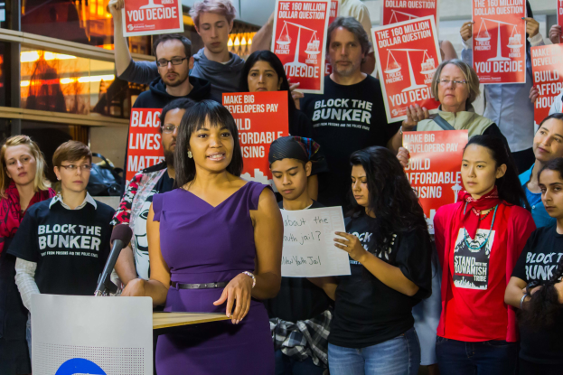 Eine Black-Lives-Matter-Aktivistin spricht an einem Podium mit Mikrofon vor einer Menge mit Schildern, mit Lichtern und anderen Gegenständen im Hintergrund.