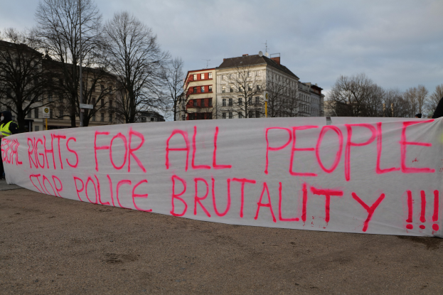 Gruppe von Menschen mit einem Banner mit der Aufschrift 'Rechte für alle Menschen Stoppt Polizeigewalt' vor einer Straßenlaterne, einem Schild, Bäumen, Gebäuden und einem bewölkten Himmel.