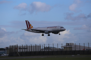 Germanwings Airbus A320-200 startet von Frankfurt Airport, mit Gras darunter, einem Zaun im Vordergrund, Gebäuden und Bäumen im Hintergrund und einer bewölkten Himmel