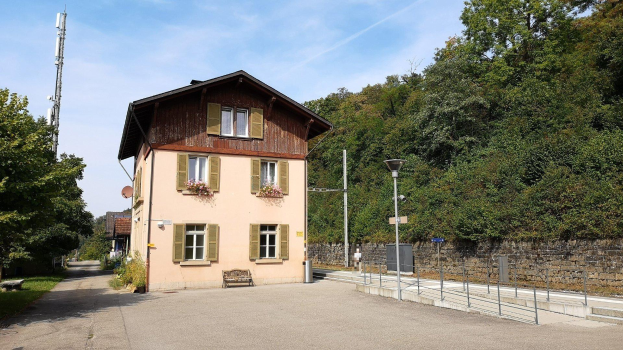 Ferienhaus zum Verkauf in Baden-Württemberg, Deutschland, mit einem Gebäude mit vielen Fenstern, einem vorderen Zaun, Straßenlaternen, Lampen, Grills, einer Bank, blühenden Pflanzen, Gras, Bäumen, einer Wand und einem bewölkten Himmel.