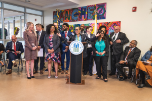Gruppe von Menschen vor einem Podium mit Mikrofon und einer Tafel mit Text, mit sitzenden und stehenden Teilnehmern, Flaggen, einem Feuerlöscher und Deckenleuchten im Hintergrund.