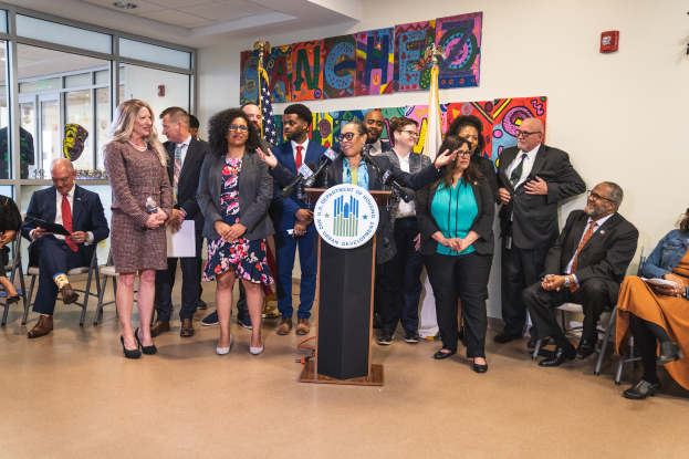 Gruppe von Menschen vor einem Podium mit Mikrofon und einer Tafel mit Text, mit sitzenden und stehenden Teilnehmern, Flaggen, einem Feuerlöscher und Deckenleuchten im Hintergrund.