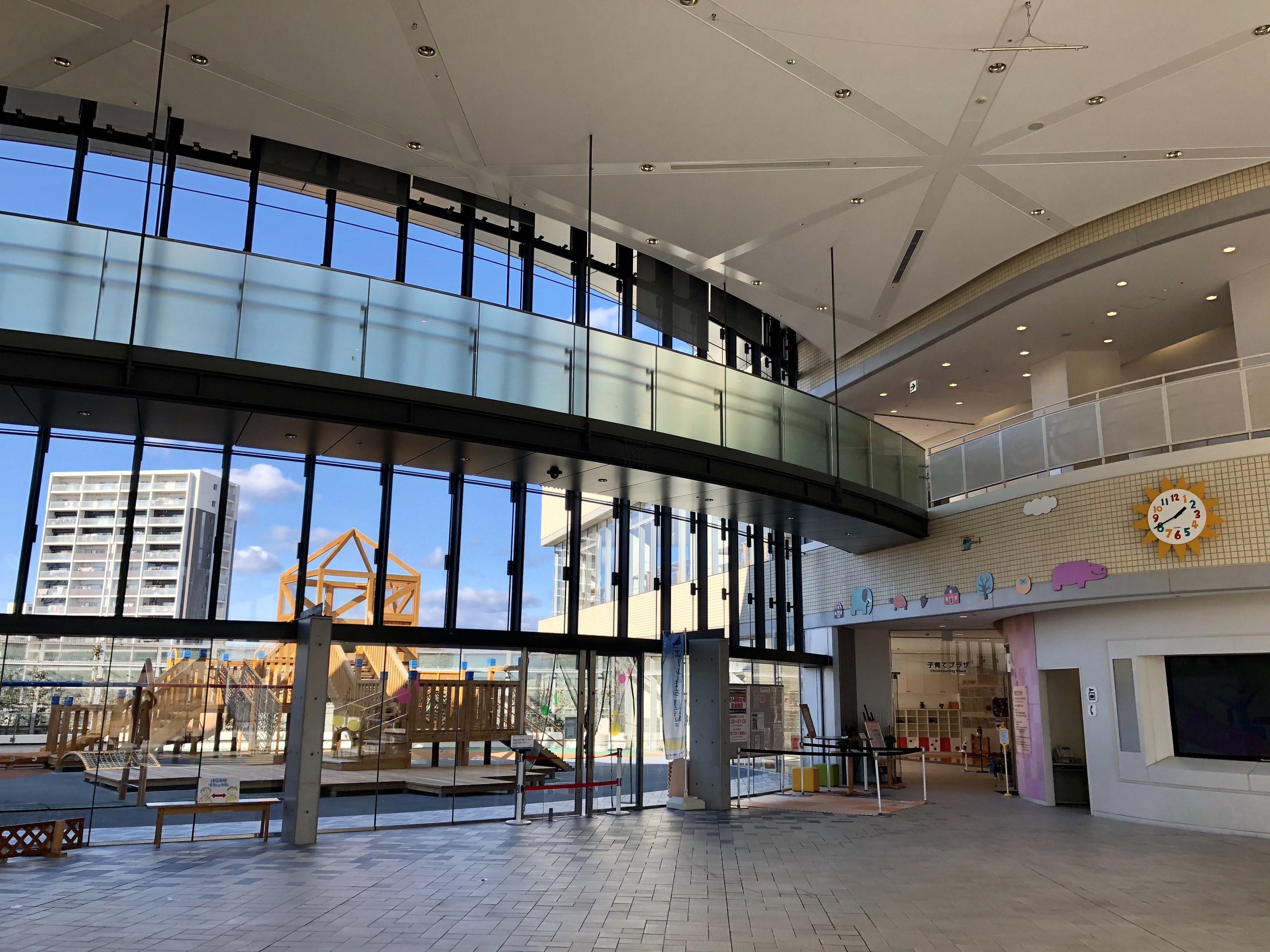 Innenansicht eines großen Gebäudes mit hohen Decken, zahlreichen Fenstern, elektrischem Licht, einer wandmontierten Uhr, einem Fernseher, einem Anzeigebildschirm, hölzernen Strukturen, einem hölzernen Zaun, umgeben von Gebäuden, Bäumen und einem bewölkten Himmel, als Haupteingang zu einem neuen Kinderspielplatz.
