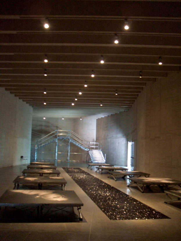 Ein geräumiger Raum im Nationalmuseum der Tschechischen Republik Holocaust-Gedenkstätte mit zahlreichen Tischen und Bänken, einem Teppichboden, Wänden, Treppen mit Gelöndern und Deckenlampen.