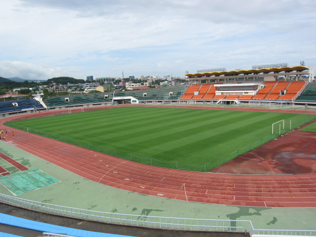 Großes Stadion mit einem Fußballfeld, umgeben von Gebäuden, Bäumen, Hügeln und einem klaren blauen Himmel, mit einigen Menschen und saftig grünem Gras.