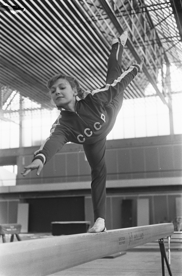 Ein junges Mädchen führt Gymnastik auf einer Stange in einer Turnhalle durch, mit einer Wand und einer Decke mit Stangen im Hintergrund.