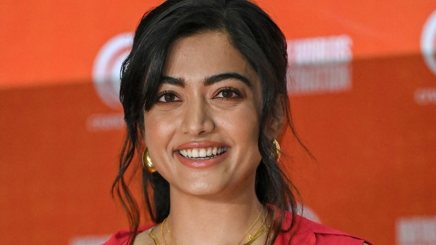 Frau in rotem Kleid mit elegantem Dutt und dezentem Make-up, die in die Kamera lächelt, vor einem gelben Banner mit der Aufschrift 'Sonam Kapoors beste Schönheits-Looks vom Cannes Film Festival'.