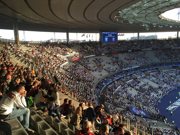 Eine große Menschenmenge sitzt im Allianz Stadion in München, Deutschland, und schaut ein Fußballspiel. Rechts im Bild ist eine Bühne mit ein paar Leuten darauf und im Hintergrund sind Fahnen, Stangen und ein Bildschirm zu sehen.