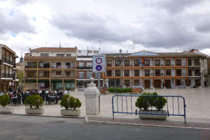 Ein belebter Stadtplatz mit sitzenden und stehenden Menschen, Topfpflanzen, einer Metallabsperrung, einem Schild an einem Pfahl, Straßenlaternen mit Fahnen, umliegenden Gebäuden mit Fenstern und einem bewölkten Himmel.