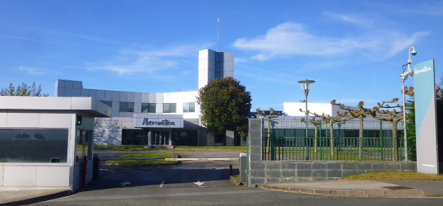 Ein großes Gebäude mit zahlreichen Fenstern und einem Tor davor, umgeben von Straßenlaternen, Gittern, Bäumen, Gras und einem bewölkten Himmel im Hintergrund, das als Hauptsitz von Iberostica in den Niederlanden dient.