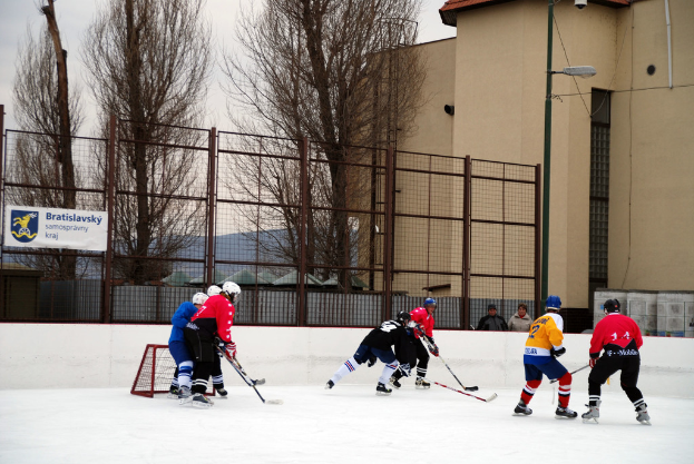 Menschen spielen Eishockey auf einer Eisfläche mit Gebäuden, Bäumen, einer Straßenlaterne, einem Namensschild und Zäunen im Hintergrund unter einem klaren Himmel.