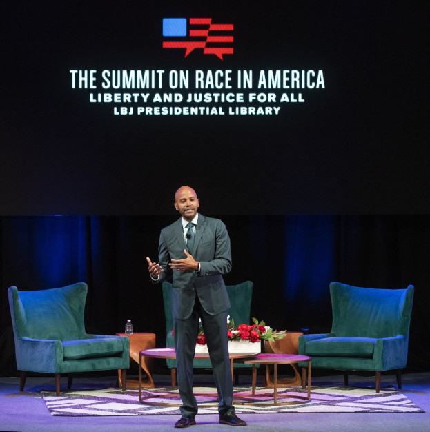 Ein Mann in einem Blazer, Krawatte und Hemd steht auf einer Bühne und spricht bei der "The Summit on Race in America: Liberty and Justice for All"-Veranstaltung in der LBJ Presidential Library, mit zwei Stühlen, einem Tisch mit einem Blumenstrauß und einer Flasche darauf und einem Bildschirm hinter ihm.