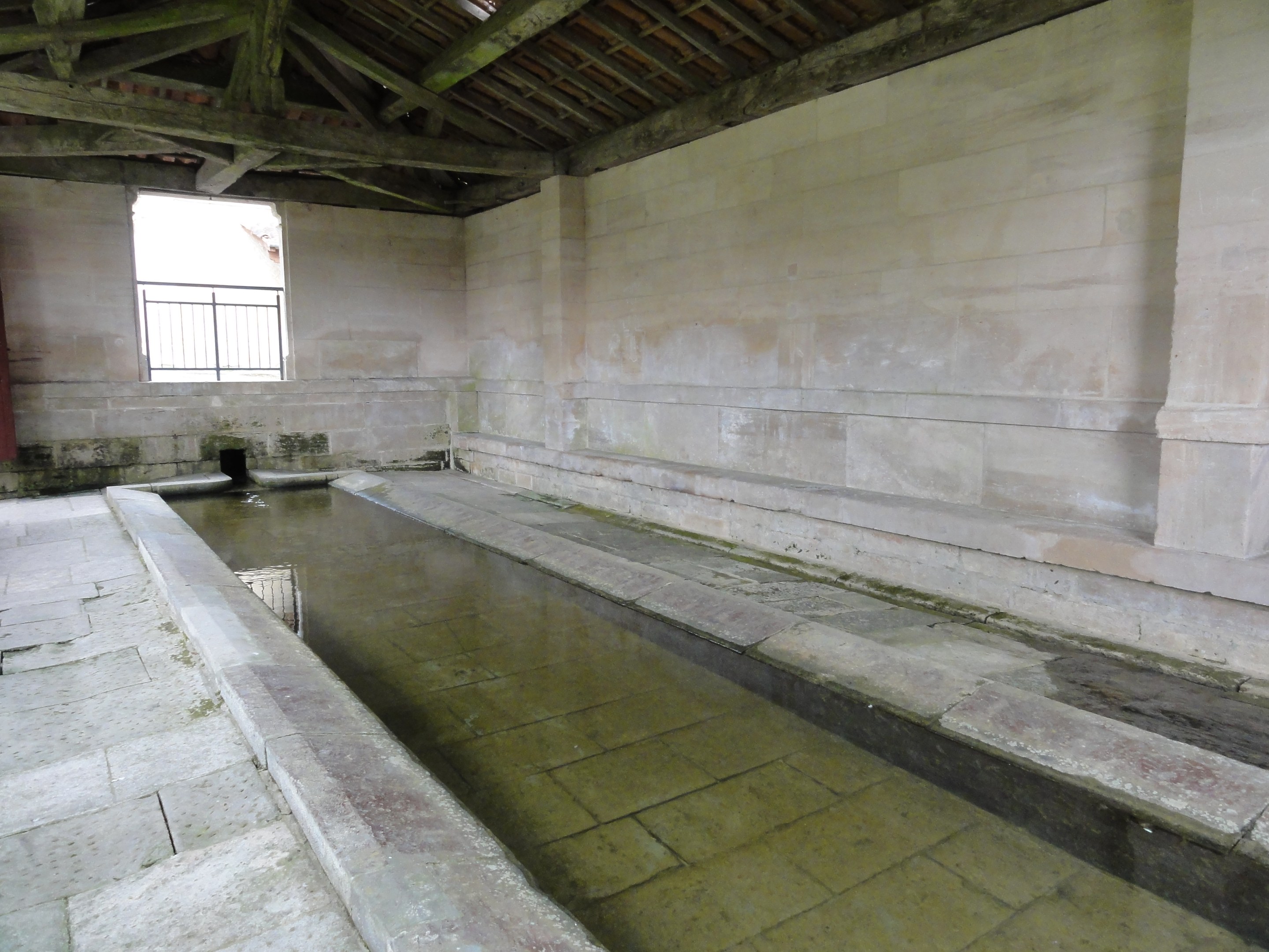 Alter Steinbau mit einem zentralen Pool, einem Fenster im Hintergrund und hölzernen Balken oben, die an eine Badeeinrichtung erinnern.