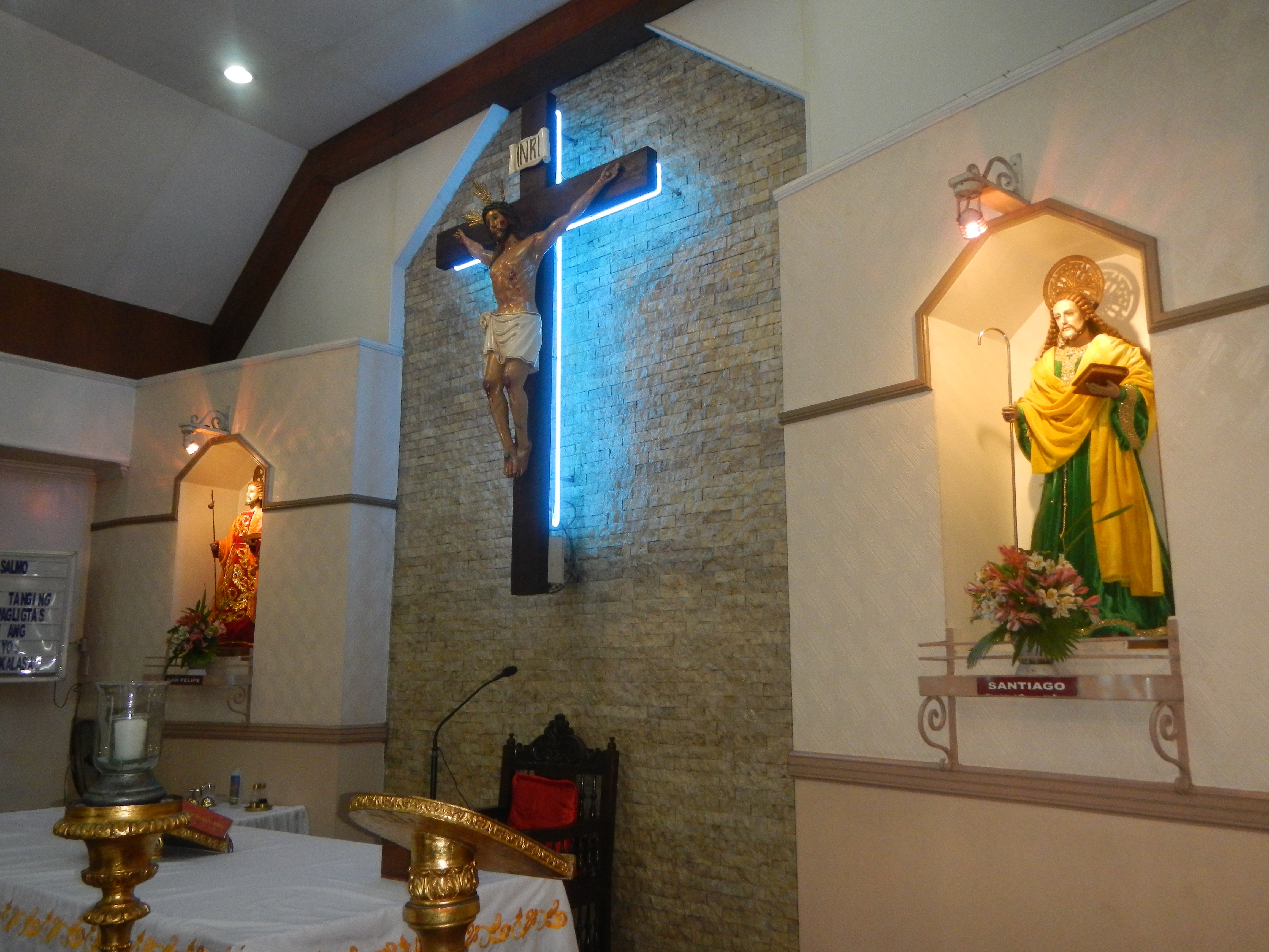 Das Innere einer Kirche mit einem Kreuz an der Wand, einer Jesus-Statue am Kreuz, einem Tisch mit einer Decke und verschiedenen Gegenständen, einem Stuhl, einem Mikrofon mit Ständer, einer Tafel mit Text links, Deckenleuchten und Blumensträußen rechts.