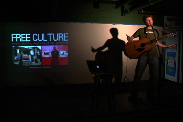Eine Person steht mit einer Gitarre in der Hand vor einem Tisch mit einem Laptop und einer Projektionsfläche im Hintergrund, auf der der Text "Free Culture" angezeigt wird.