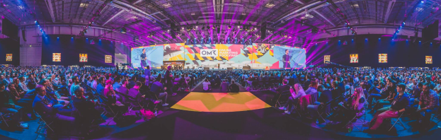 Eine große Menschenmenge sitzt vor einer hell erleuchteten Bühne mit mehreren stehenden Personen, wahrscheinlich auf einem Online-Marketing-Rockstars-Event.