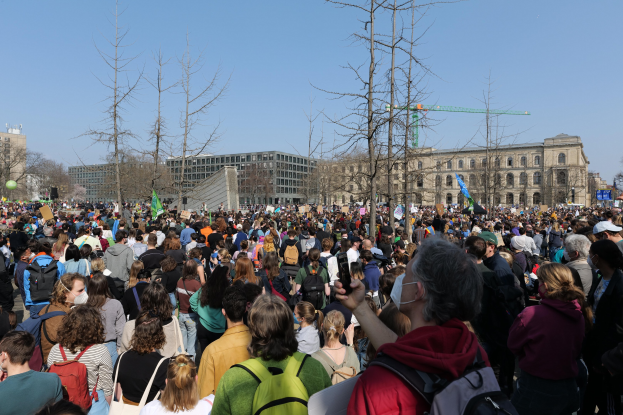 Eine große Menge von Menschen mit Schildern und Töten steht vor einem Gebäude mit Fenstern, Bäumen und einem klaren Himmel, was auf eine Klimawandel-Demonstration hindeutet.