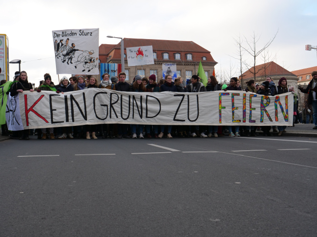Menschen mit einem Transparent mit der Aufschrift "Kein Grund zu Feiern" protestieren gegen deutsche Sparmaßnahmen auf einer Stadtstraße mit Gebäuden, Bäumen und einem klaren Himmel.