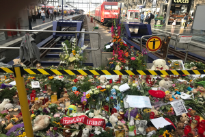 Ein Bahnsteig mit einem Zug im Hintergrund, umgeben von Menschen und verschiedenen Gegenständen wie Teddybären, Blumensträußen und Kerzen, die an einen tragischen Zugunglück in Berlin, Deutschland, erinnern.